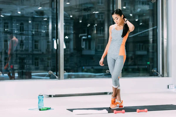 Beautiful asian sportswoman touching hair and standing on fitness mat in sports center — Stock Photo