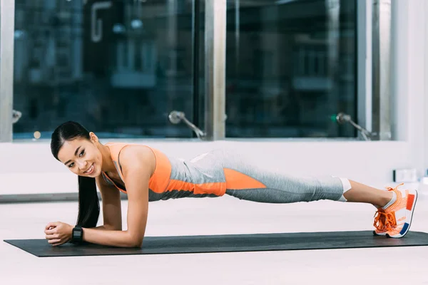 Delgado asiático sportswoman haciendo tablón en fitness mat y sonriendo en gimnasio - foto de stock