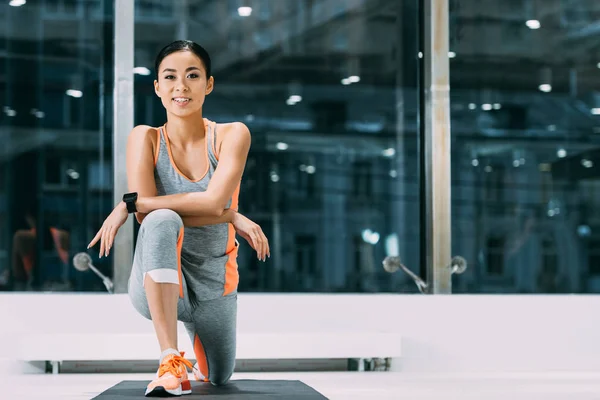 Bela asiática esportista sorrindo e fazendo exercício de alongamento no tapete de fitness no centro de esportes — Fotografia de Stock