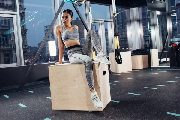 Attractive asian sportswoman sitting on big wooden cube in sports center — Stock Photo