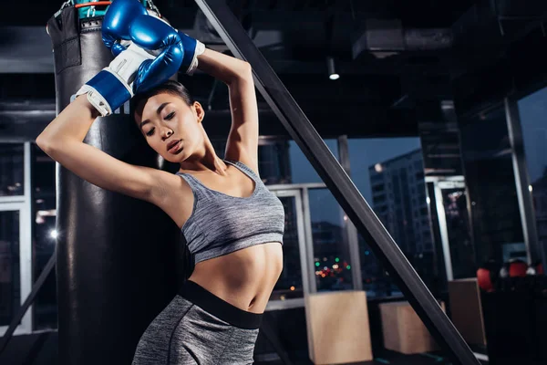 Привлекательная сексуальная азиатская спортсменка в боксёрских перчатках, растянувшаяся рядом с боксёрской грушей в спортзале — стоковое фото