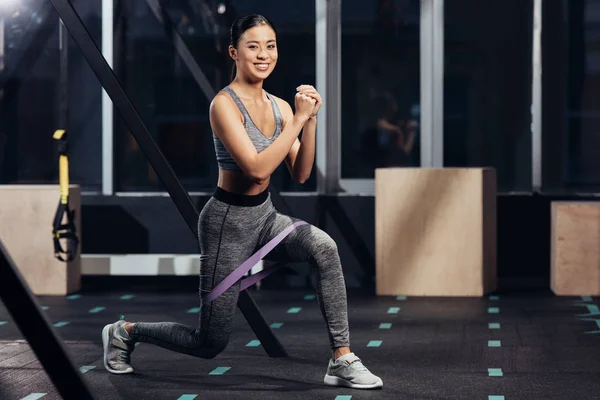 Sorridente ragazza asiatica che fa affondi con elastici di resistenza in palestra — Foto stock