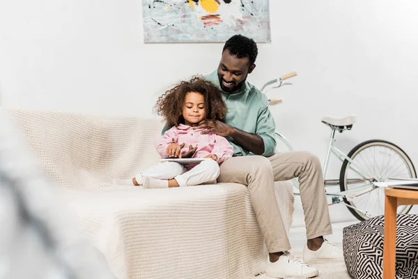 Padre e hija afroamericanos cómodamente sentados en el sofá y usando tableta digital - foto de stock