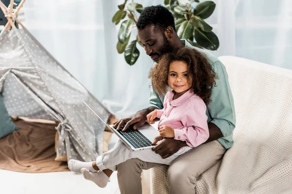 Africano-americano padre y el niño usando el ordenador portátil mientras está sentado en el sofá cerca de wigwam - foto de stock