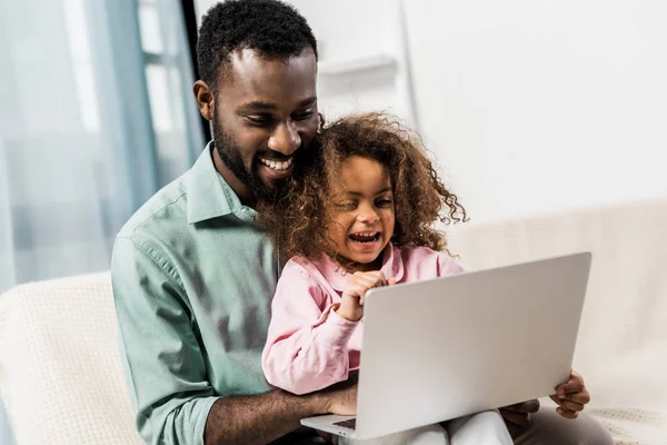 Niño afroamericano sonriente sentado con el padre en el sofá y usando el ordenador portátil - foto de stock