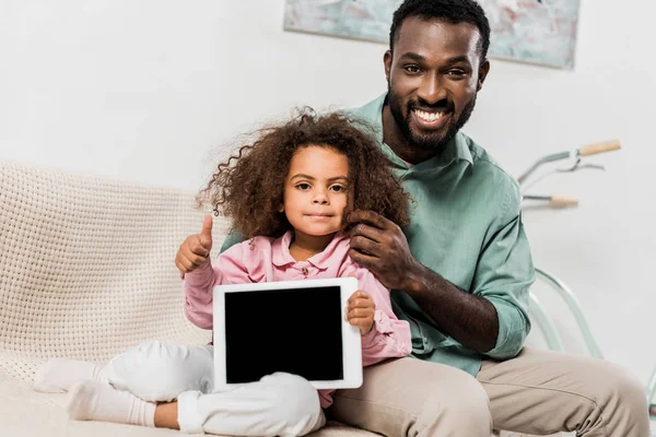 Africano americano padre y hija sentado en sofá con digital tablet y mirando a cámara - foto de stock