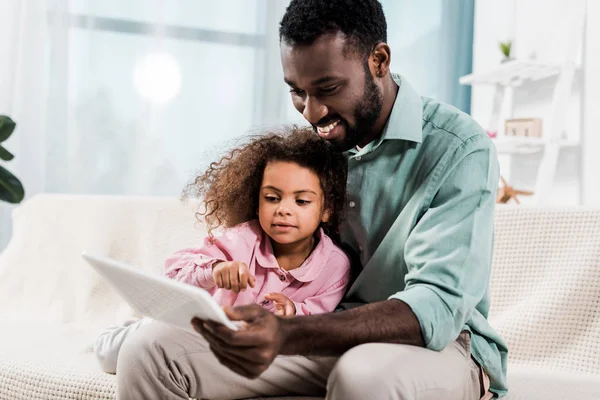 Beau père afro-américain et sa fille en utilisant une tablette numérique tout en étant assis sur le canapé — Photo de stock