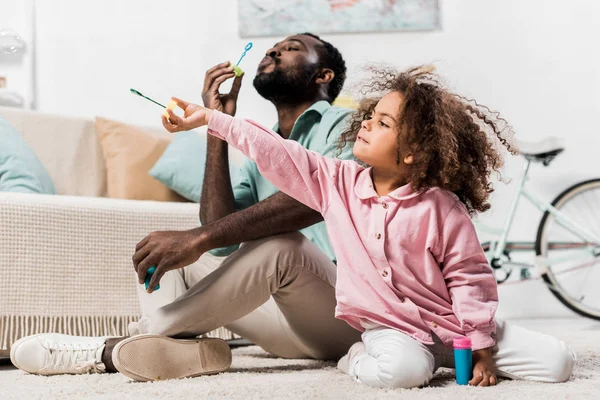 Familia afroamericana soplando burbujas de jabón en la sala de estar - foto de stock