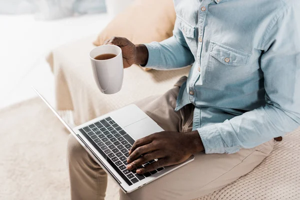 Africano americano freelancer segurando xícara de chá enquanto digita no laptop e sentado no sofá — Fotografia de Stock