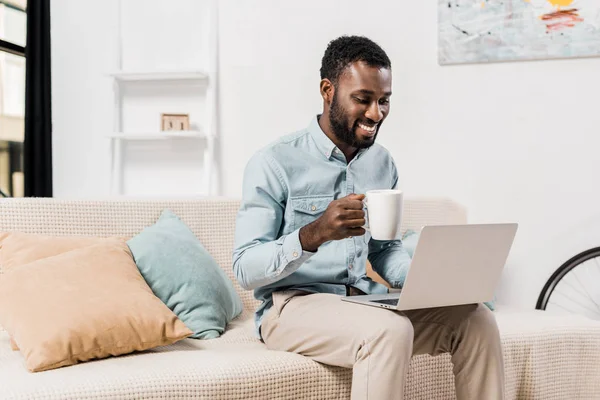 Sonriente afroamericano freelancer beber té y el uso de ordenador portátil en la sala de estar - foto de stock