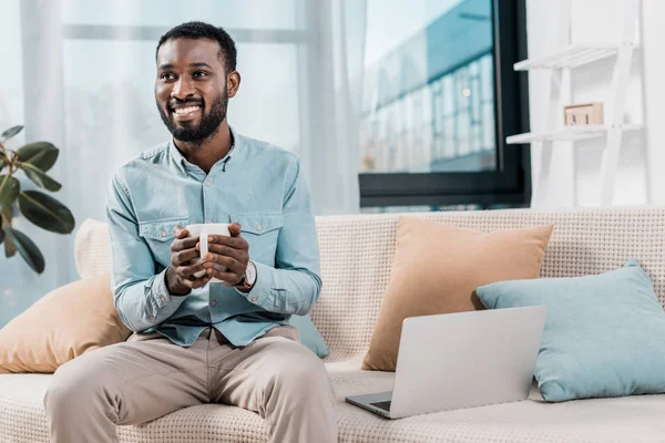 Freelance afroamericano sorridente mentre tiene in mano una tazza di tè e si siede sul divano in soggiorno — Foto stock