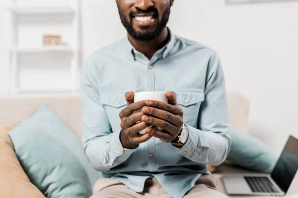 Vue recadrée d'un homme afro-américain souriant tenant une tasse de thé et assis sur un canapé — Photo de stock