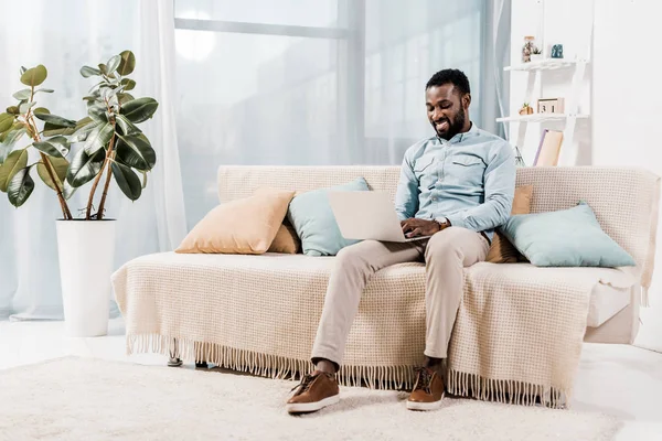 Africano americano freelancer sentado no sofá na sala de estar, usando laptop — Fotografia de Stock