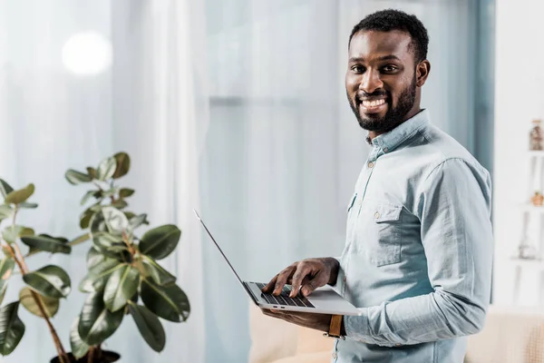 Freelancer afroamericano sonriendo y sosteniendo laptop - foto de stock