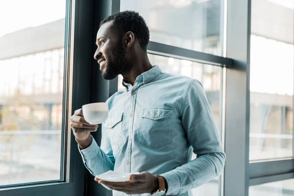 Uomo afroamericano sorridente che tiene in mano una tazza di caffè e guarda fuori dalla finestra — Foto stock