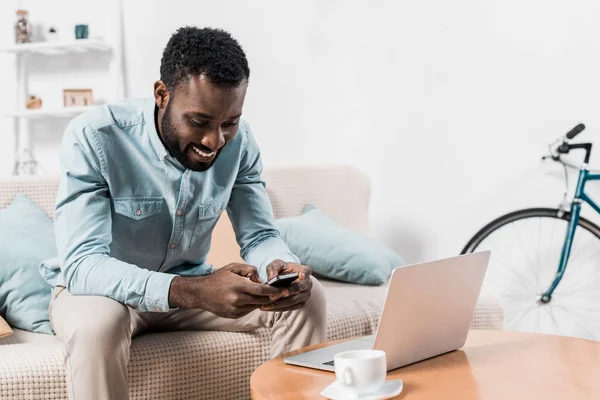 Африканский американский фрилансер сидит на диване и печатает на смартфоне — стоковое фото