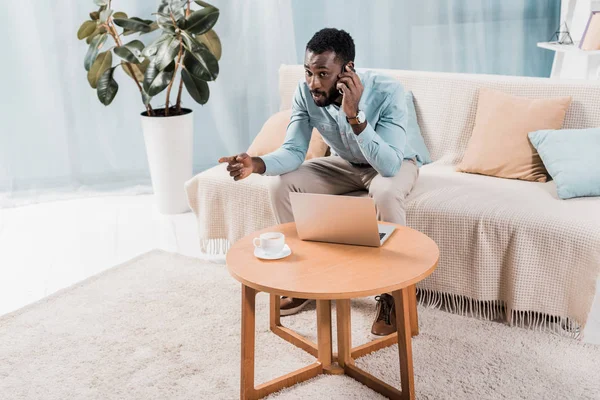 Beau freelance afro-américain assis sur le canapé et parlant sur smartphone — Photo de stock
