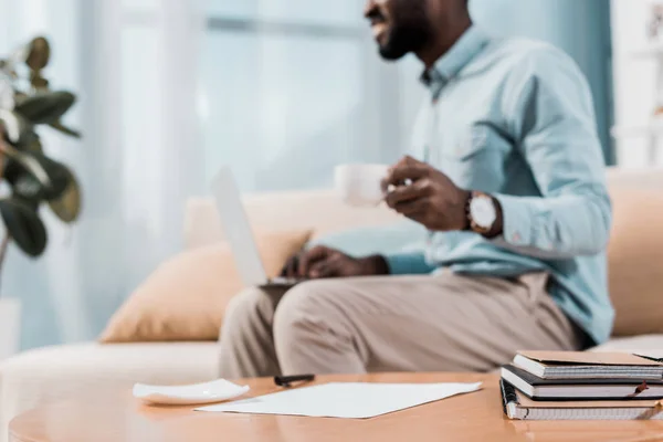 Abgeschnittene Ansicht eines afrikanisch-amerikanischen Freelancers, der mit Laptop auf der Couch sitzt und Kaffee trinkt — Stockfoto