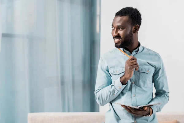 Sorrindo afro-americano com notebook e lápis desviando o olhar — Fotografia de Stock