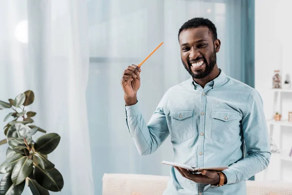 Sonriente afroamericano freelancer sosteniendo bloc de notas y lápiz mientras mira a la cámara - foto de stock