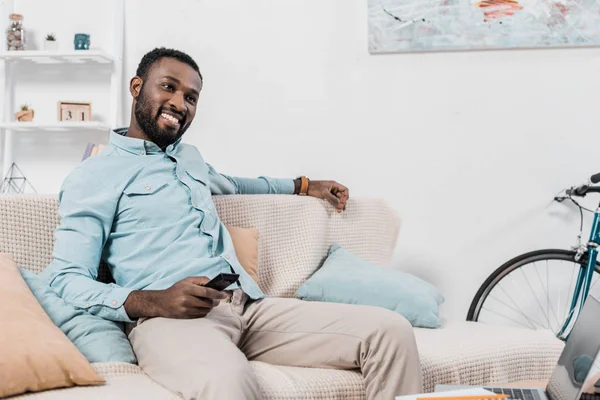 Feliz homem americano africano sentado no sofá com controlador remoto — Fotografia de Stock