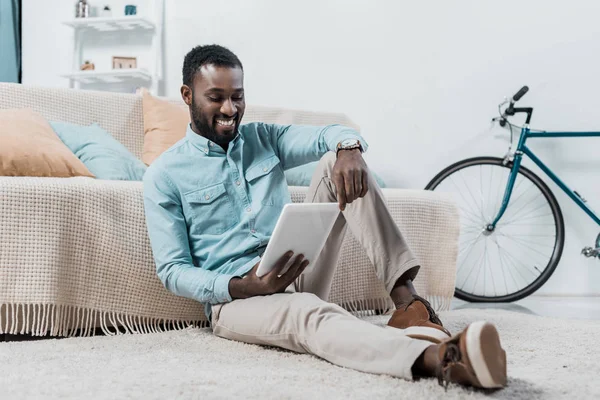 Homme afro-américain travaillant sur tablette numérique et assis sur le sol dans le salon — Photo de stock