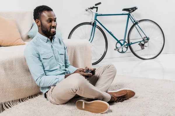 Homme afro-américain assis sur le sol et jouer à un jeu vidéo avec vélo bleu sur fond — Photo de stock