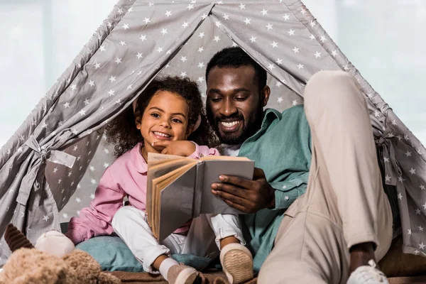 Padre afroamericano leyendo libro con niño y sonriendo en la sala de estar - foto de stock