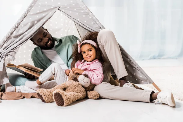 Африканский американский отец читает книгу с ребенком и улыбается в wigwam — стоковое фото