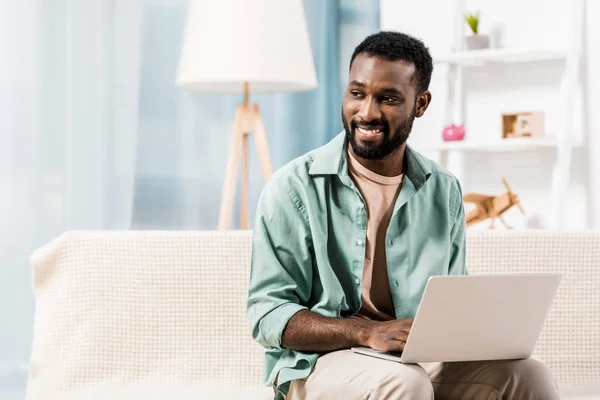 African american man using laptop and looking away in living room — Stock Photo