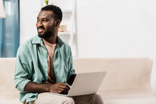 Афроамериканець людиною, використовуючи ноутбук на колінах і фотографіях хтось дивитися вбік у вітальні — стокове фото