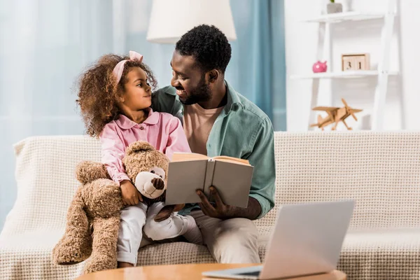 Papá afroamericano leyendo con su hija y mirándose en la sala de estar - foto de stock