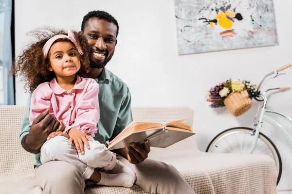 Afroamericano padre leyendo con hija sentado de rodillas en sala de estar - foto de stock