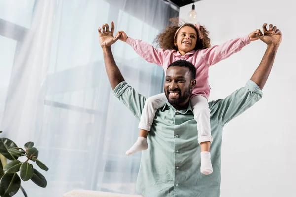 Afro-americano pai carregando filha no pescoço e brincando na sala de estar — Fotografia de Stock