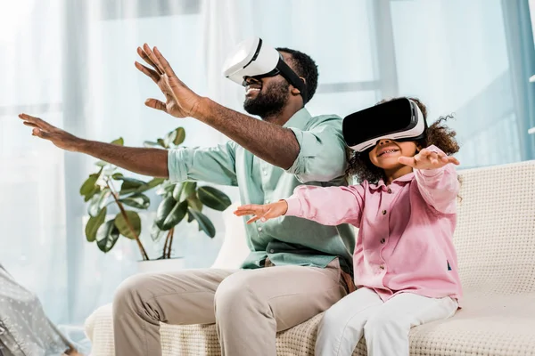 Padre e hija afroamericanos usando gafas de realidad virtual y sonriendo en la sala de estar - foto de stock