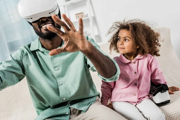 Hombre afroamericano con gafas de realidad virtual e hija sentada en sofá - foto de stock