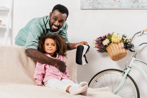 Afro-americano pai abraçando e mostrando filha virtual realidade óculos na sala de estar — Fotografia de Stock