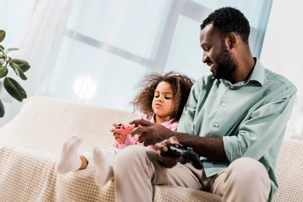 African american dad and daughter sitting on sofa and playing video game in living room — Stock Photo