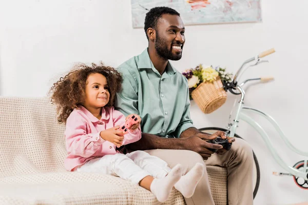 Padre e hija afroamericanos sentados en el sofá y jugando videojuegos juntos en la sala de estar - foto de stock