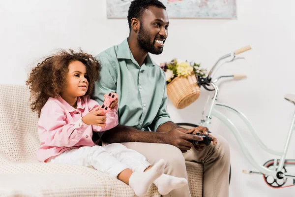 Padre e hija afroamericanos jugando videojuegos en la sala de estar - foto de stock