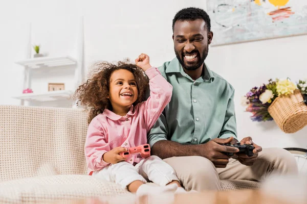 Padre afroamericano y su hija jugando videojuegos con joysticks en la sala de estar - foto de stock