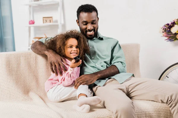Афроамериканський тато обіймає дочку під час перегляду телевізора у вітальні — стокове фото