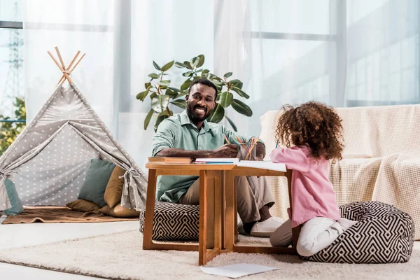 African american dad drawing with daughter and looking at camera in living room — Stock Photo