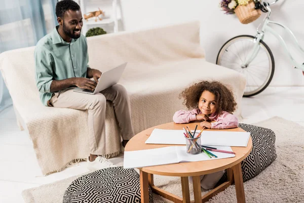 Homem americano africano trabalhando no laptop e filha desenho com lápis na sala de estar — Fotografia de Stock