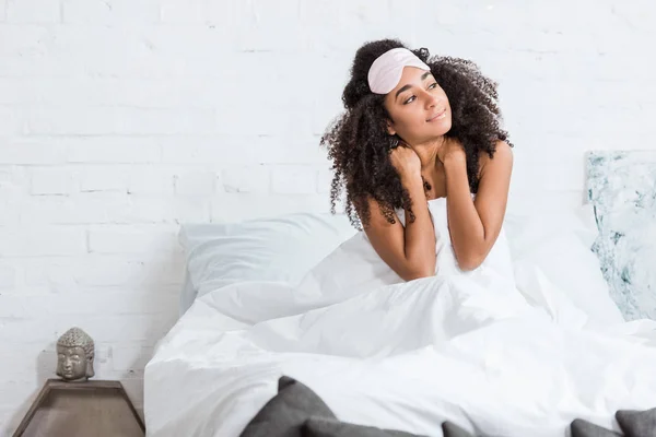 Mujer afroamericana con los ojos vendados en la frente sentada en la cama durante la mañana en casa - foto de stock