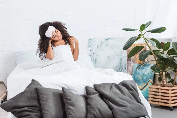 Heureuse jeune femme afro-américaine avec les yeux bandés sur le front assis au lit le matin à la maison — Photo de stock