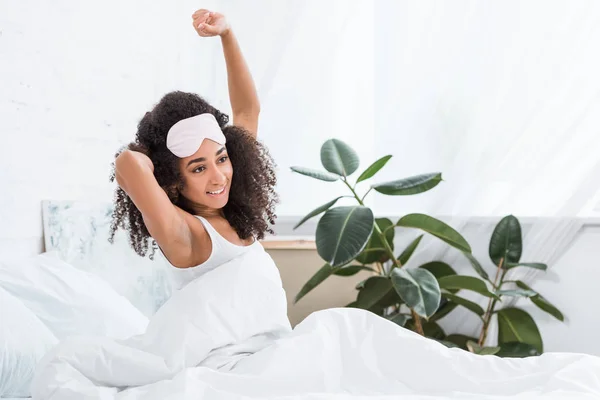 Alegre joven afroamericana con los ojos vendados en la frente estirándose en la cama durante la mañana en casa - foto de stock