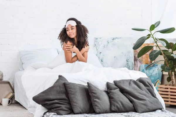 Mujer afroamericana bonita con los ojos vendados en la frente sentada en la cama durante la mañana en casa - foto de stock