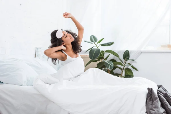 Vista lateral de la joven afroamericana con los ojos vendados en la frente estirándose en la cama durante la mañana en casa - foto de stock