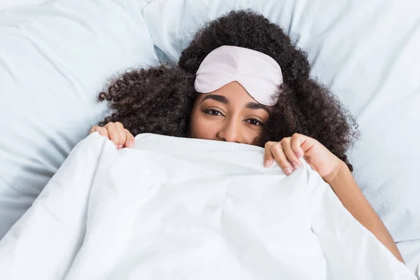 Happy young african american woman with blindfold on forehead covering face by blanket and laying in bed during morning time at home — Stock Photo